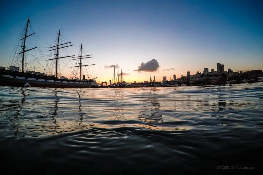 View from the water at Aquatic Park in San Francisco showing the historic sailing ship Balclutha at sunrise, with rippling bay waters and city skyline in the background.