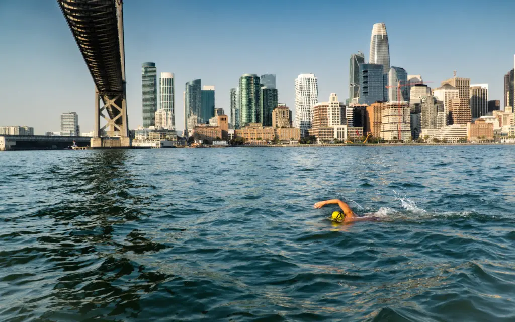 Swimmer in a yellow cap moves through San Francisco Bay beneath the Bay Bridge, with the city skyline visible in the background