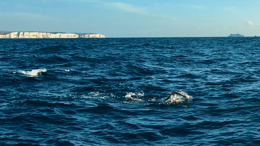 Swimmer Sylvia Lacock crossing the English Channel in open water, with the White Cliffs of Dover in the background—an image capturing the moment before a swim-ending injury that later inspired the founding of Pacific Open Water Swim Co.